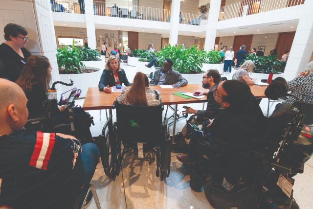 A group of people in wheelchairs and standing are gathered around a large table in a spacious, well-lit atrium filled with plants. They appear to be engaged in a discussion or meeting, with some individuals using laptops and notebooks. The atmosphere is collaborative and inclusive, featuring a diverse group of attendees.