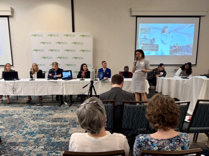 A conference setting featuring a panel of speakers at tables equipped with microphones and laptops. In the foreground, an audience is seated, facing a woman in a white dress speaking into a microphone. The backdrop displays a presentation screen with images. The environment appears professional, with a neutral color scheme and branding visible on the walls.