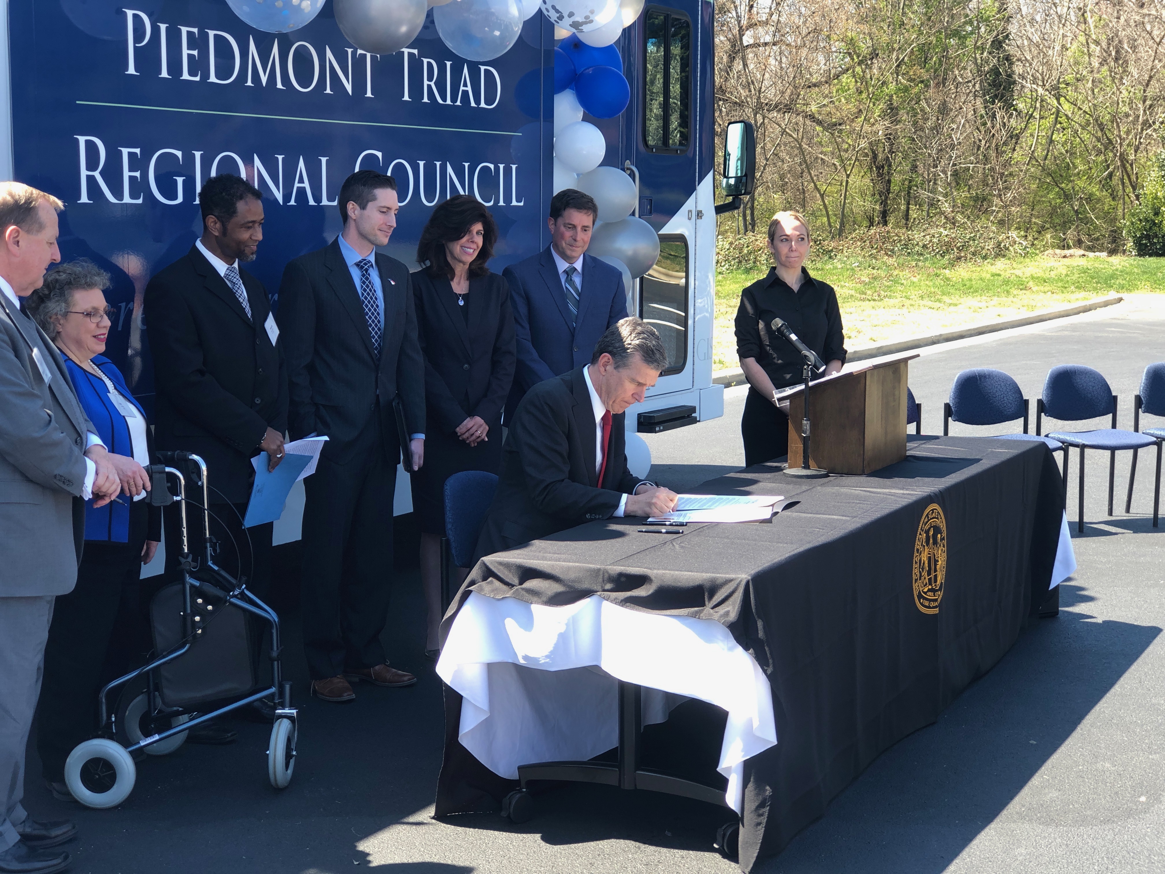 Governor Roy Cooper is at a desk signing Employment First Executive Order. Behind him are leaders from NC DHHS, NC Vocational Rehabilitation and NCCDD.